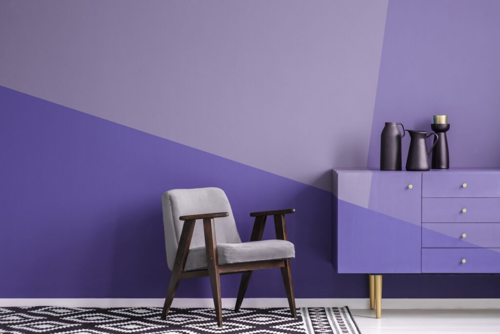 Real photo of a gray, wooden armchair on patterned, black and white rug in creative living room interior with geometric, violet wall and cupboard Color Blocking Interior Design